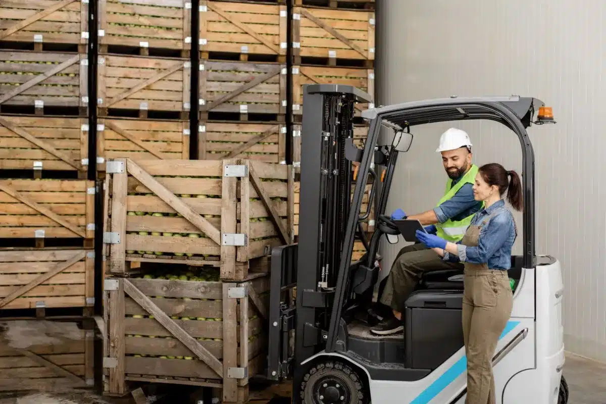 Two warehouse workers operating forklift near stacked wooden crates in logistics facility