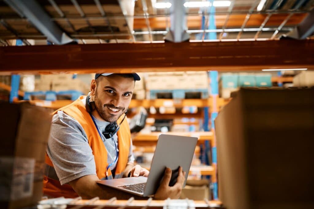 warehouse worker holding a laptop and looking through a warehouse shelf